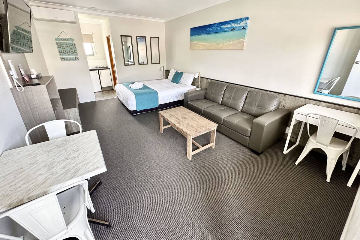 A hotel room with a bed, a sofa, a desk, and a chair. There is a painting on the wall and a mirror above the desk.