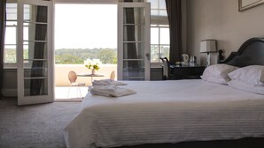 Ein Hotelzimmer mit einem großen Bett, einem Schreibtisch mit Stuhl, einem Tisch mit Blumenvase und Blick ins Freie durch offene Türen.