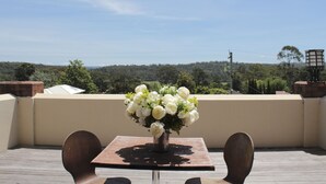 Eine Terrasse mit Tisch und Stühlen, einer weißen Blumenvase und Blick auf eine bewaldete Landschaft.