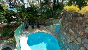 A swimming pool with a stone wall and a glass railing, surrounded by greenery and a staircase. - Little Cove Court (Noosa Heads)