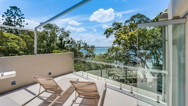 A balcony with two lounge chairs and a glass railing overlooking a scenic view of trees and the sea. - Little Cove Court (Noosa Heads)