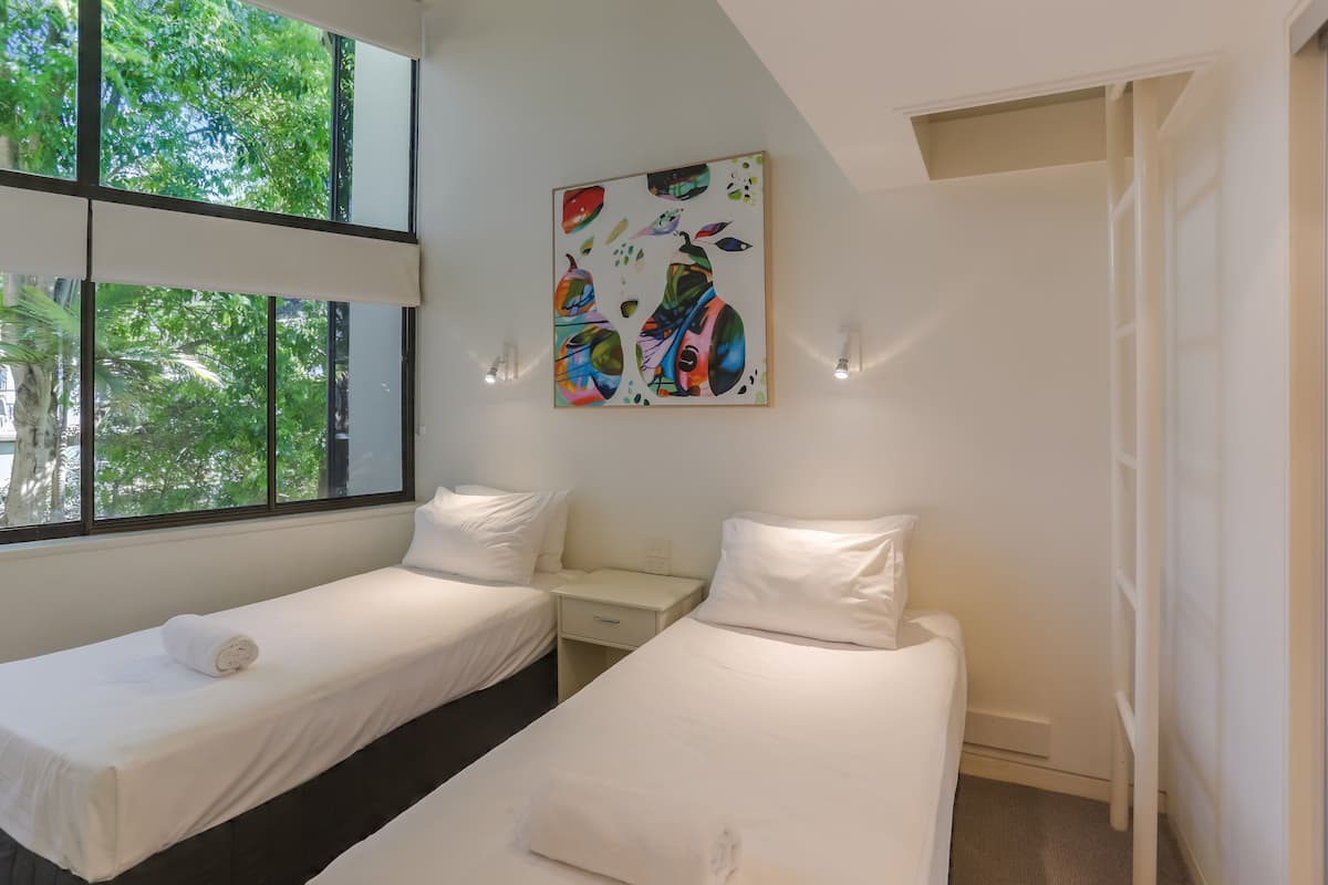A hotel room with two single beds, a small bedside table, a wall-mounted painting, and large windows offering a view of trees.