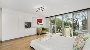 A modern bedroom with a large bed, a television, a sofa, and a balcony with a view of trees. - Little Cove Court (Noosa Heads)
