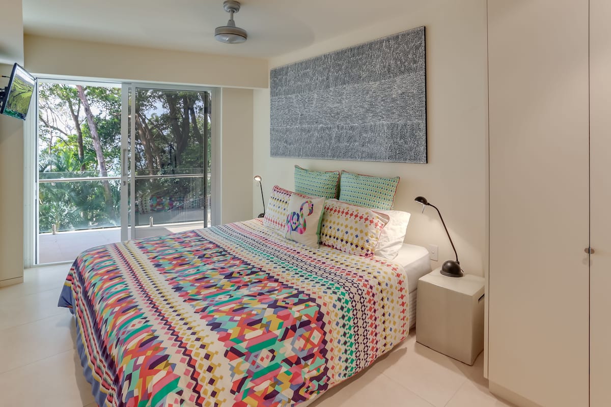 A bedroom with a large bed, a view of trees through a sliding door, and a ceiling fan.