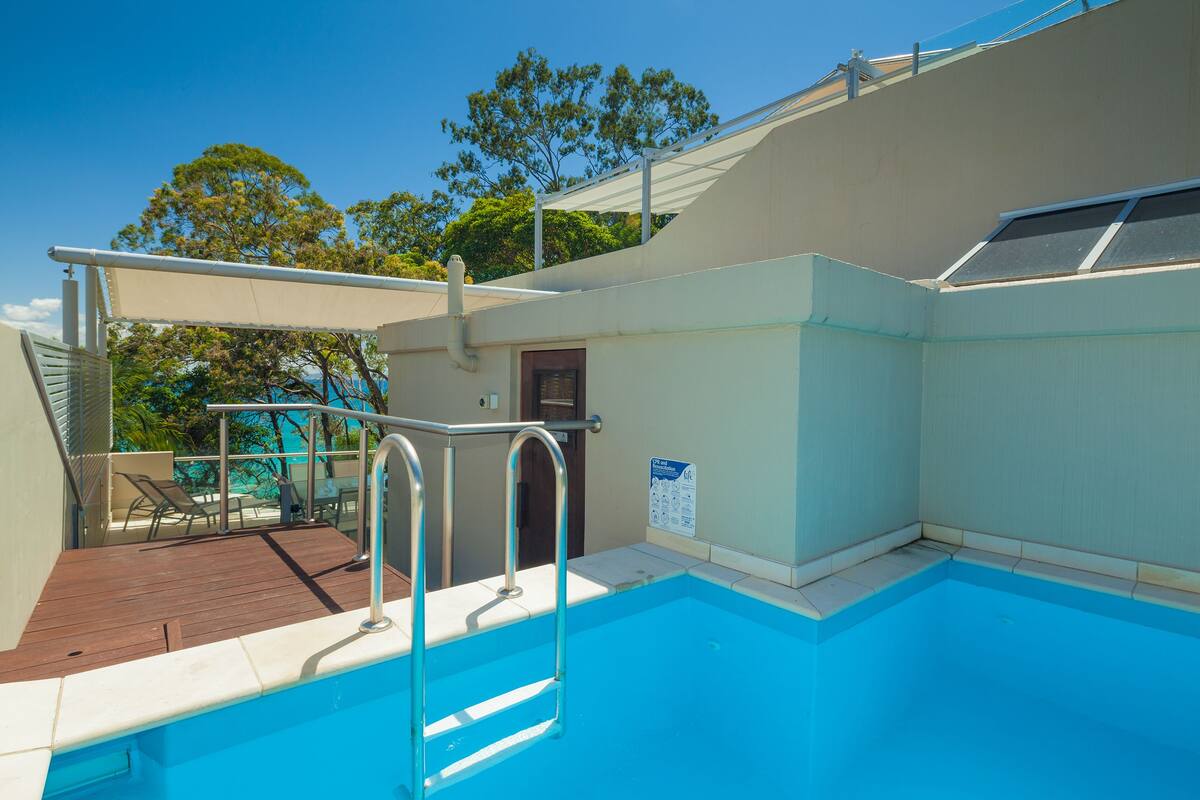 A swimming pool with a wooden deck and metal ladder, surrounded by a covered area with chairs.