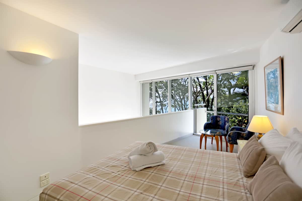 A bedroom with a large bed, a window with a view of trees, a chair, and a lamp.