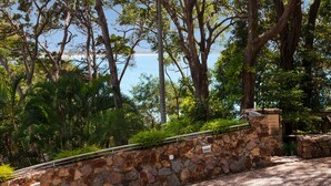A stone wall with a metal railing, surrounded by lush greenery and trees, overlooking a body of water. - Little Cove Court (Noosa Heads)