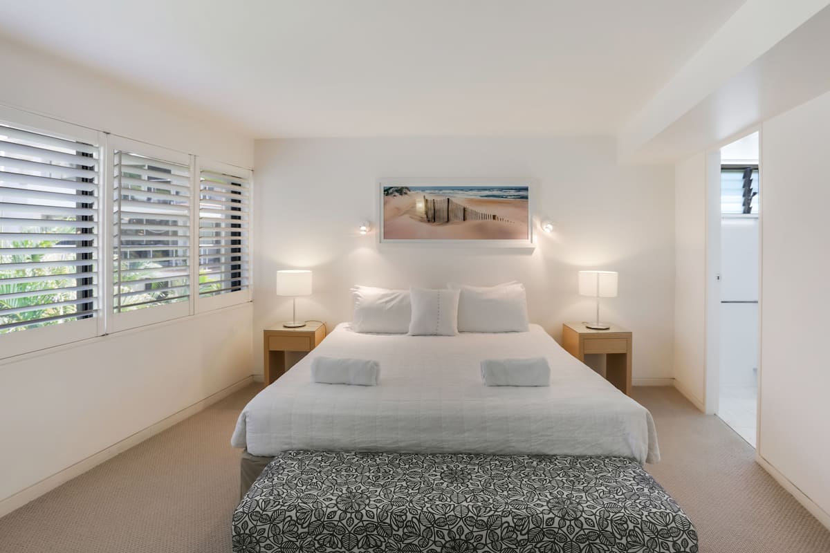 A hotel room with a large bed, two bedside tables with lamps, a painting above the bed, and a window with blinds.