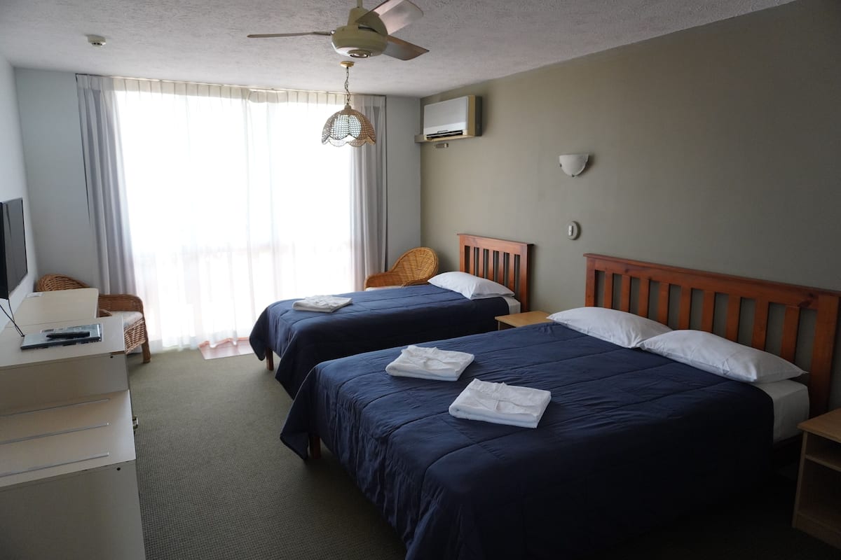 Une chambre d’hôtel avec deux lits, un bureau, une chaise, un ventilateur de plafond et une fenêtre avec des rideaux.