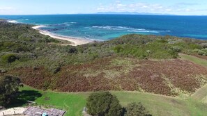 An aerial view of a coastal area with a beach, dense forest, and a clear view of the ocean.