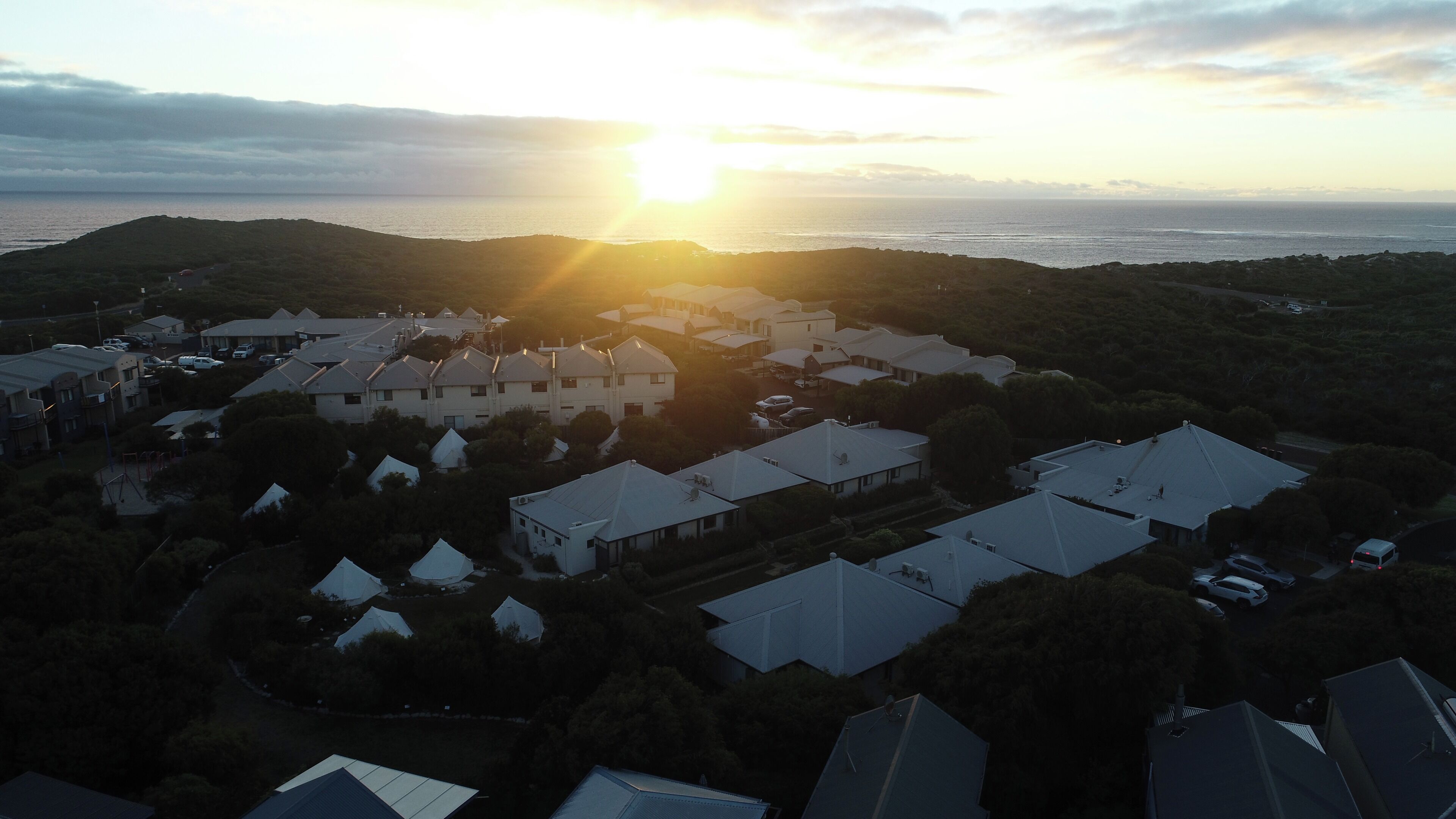 Un quartier résidentiel comprenant plusieurs maisons, une aire de jeux et offrant une vue sur la mer au coucher du soleil.