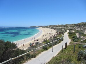 A coastal road with a beach on the right, a clear blue sky, and a few people on the beach.