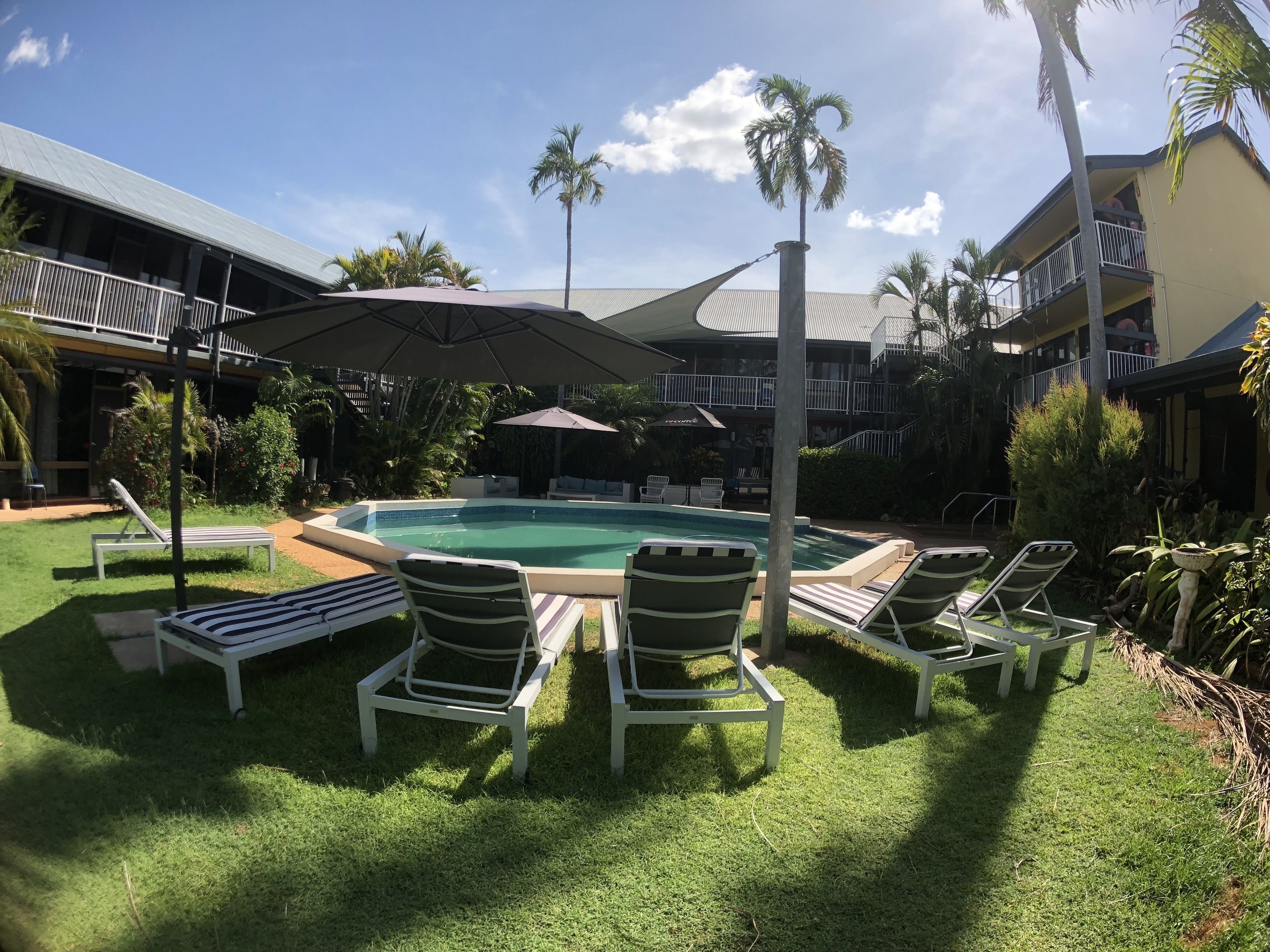 A pool area with lounge chairs and umbrellas, surrounded by greenery and a building with balconies.