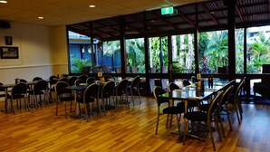 Un vaste espace de restauration intérieur doté d’un plancher en bois, de nombreuses tables et chaises, et de grandes fenêtres offrant une vue sur une végétation luxuriante.