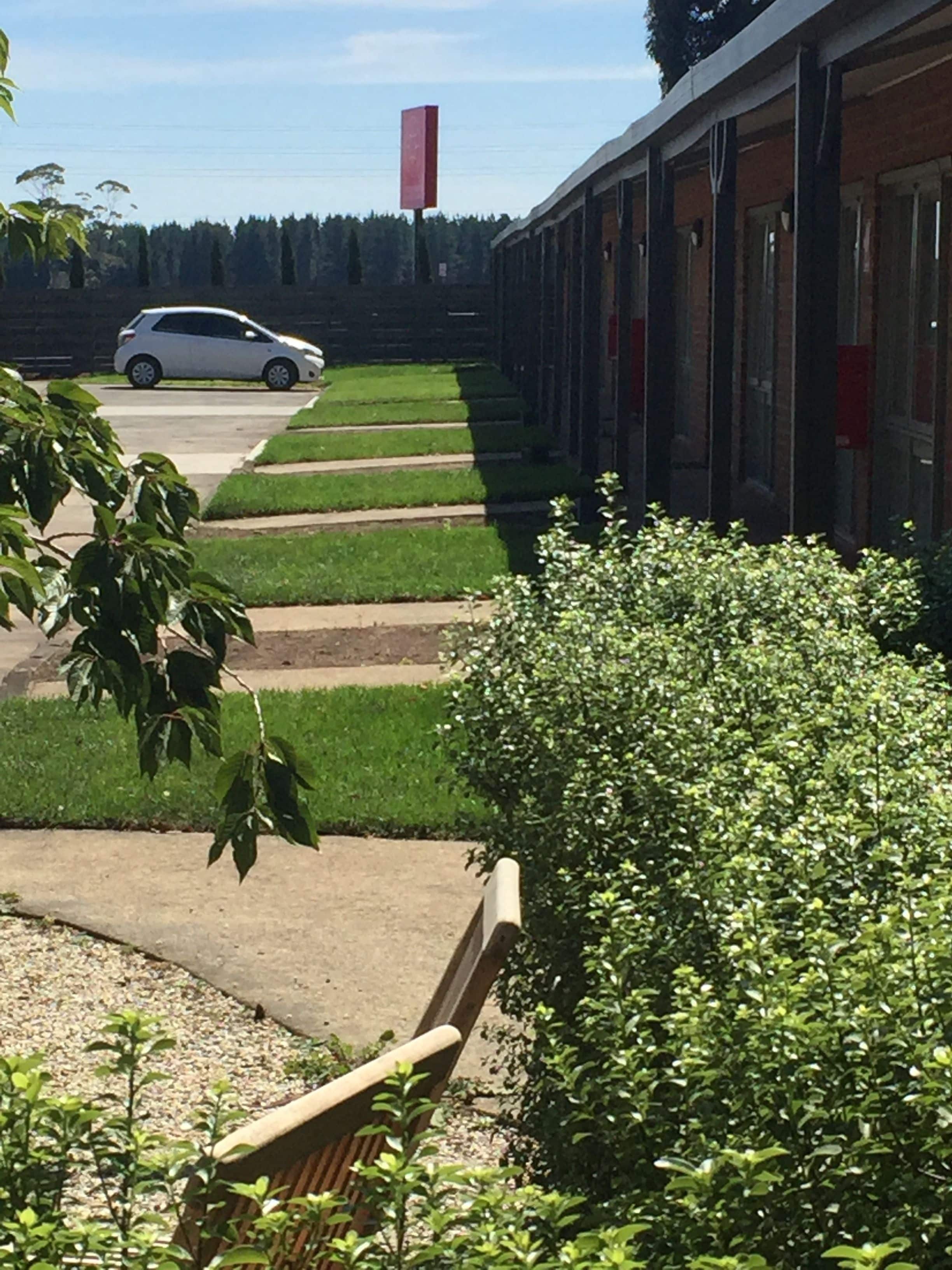 A row of single-story buildings with a car parked in front. There are green lawns and a bench in the foreground.