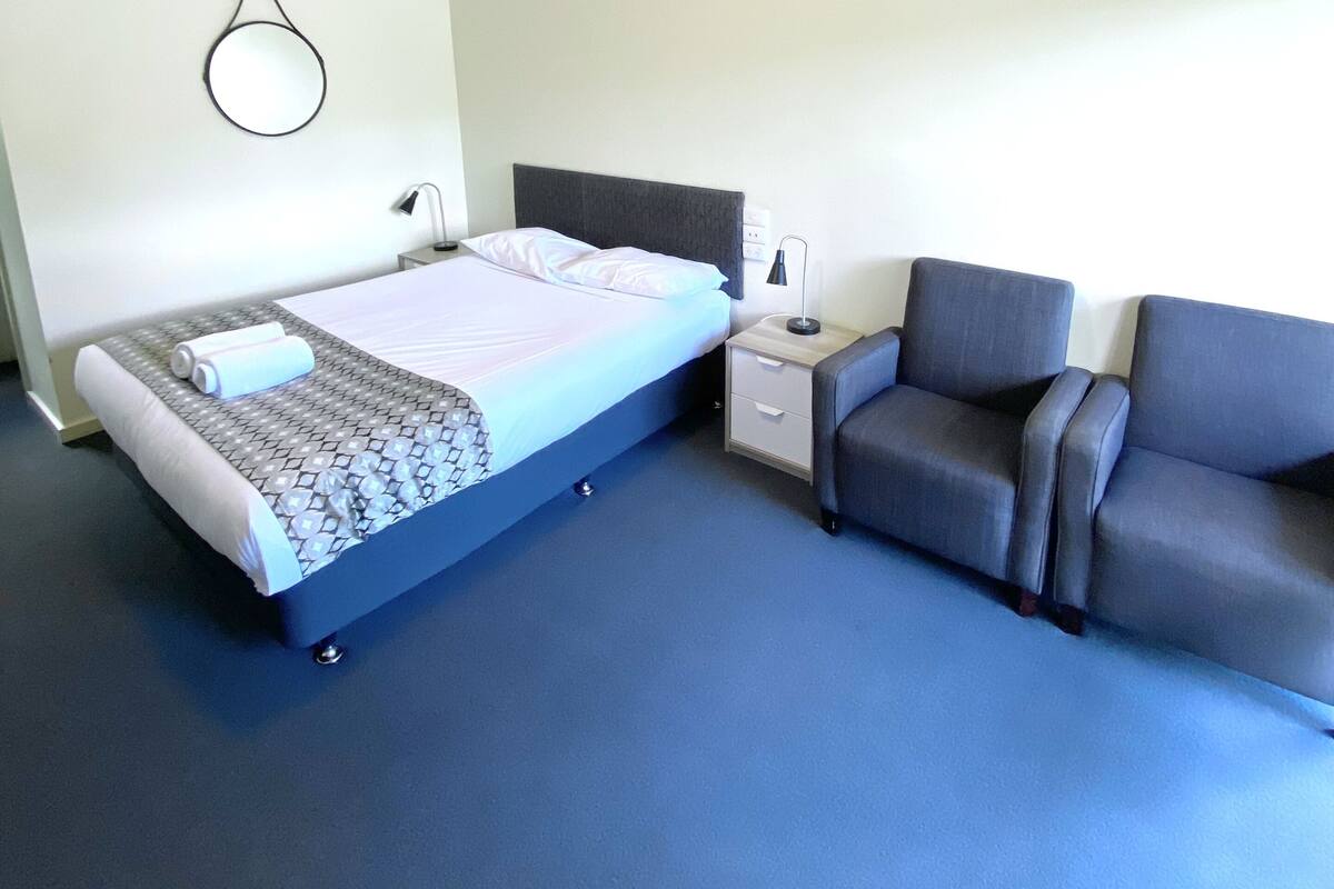 A hotel room with a bed, a nightstand, a lamp, and a gray armchair.