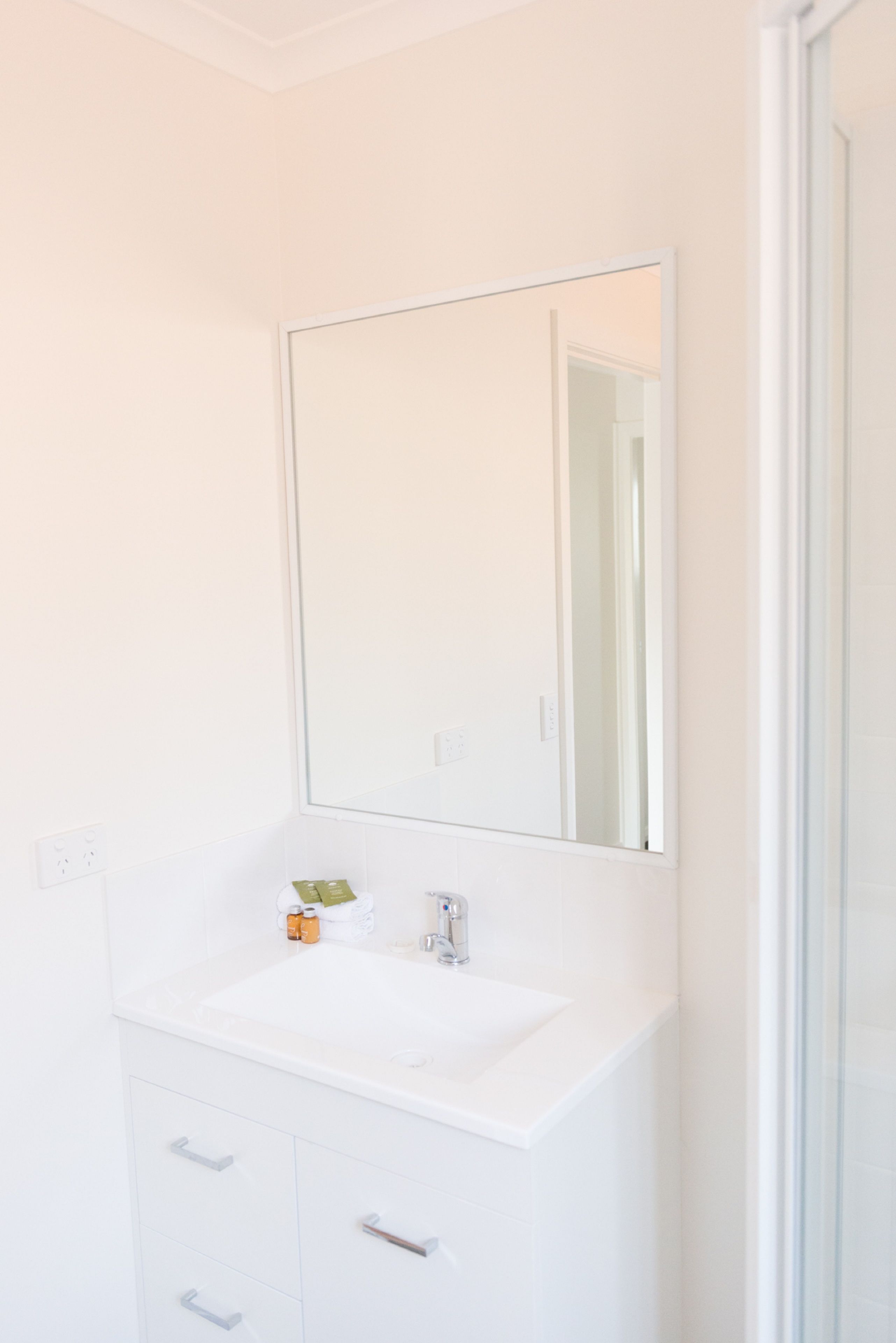 Une salle de bain blanche avec un grand miroir, un lavabo blanc et un meuble avec des tiroirs.