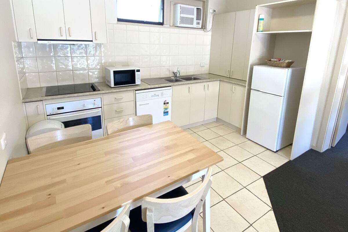 A compact kitchen with white cabinets, a microwave, a dishwasher, and a refrigerator. There is a wooden dining table with chairs.