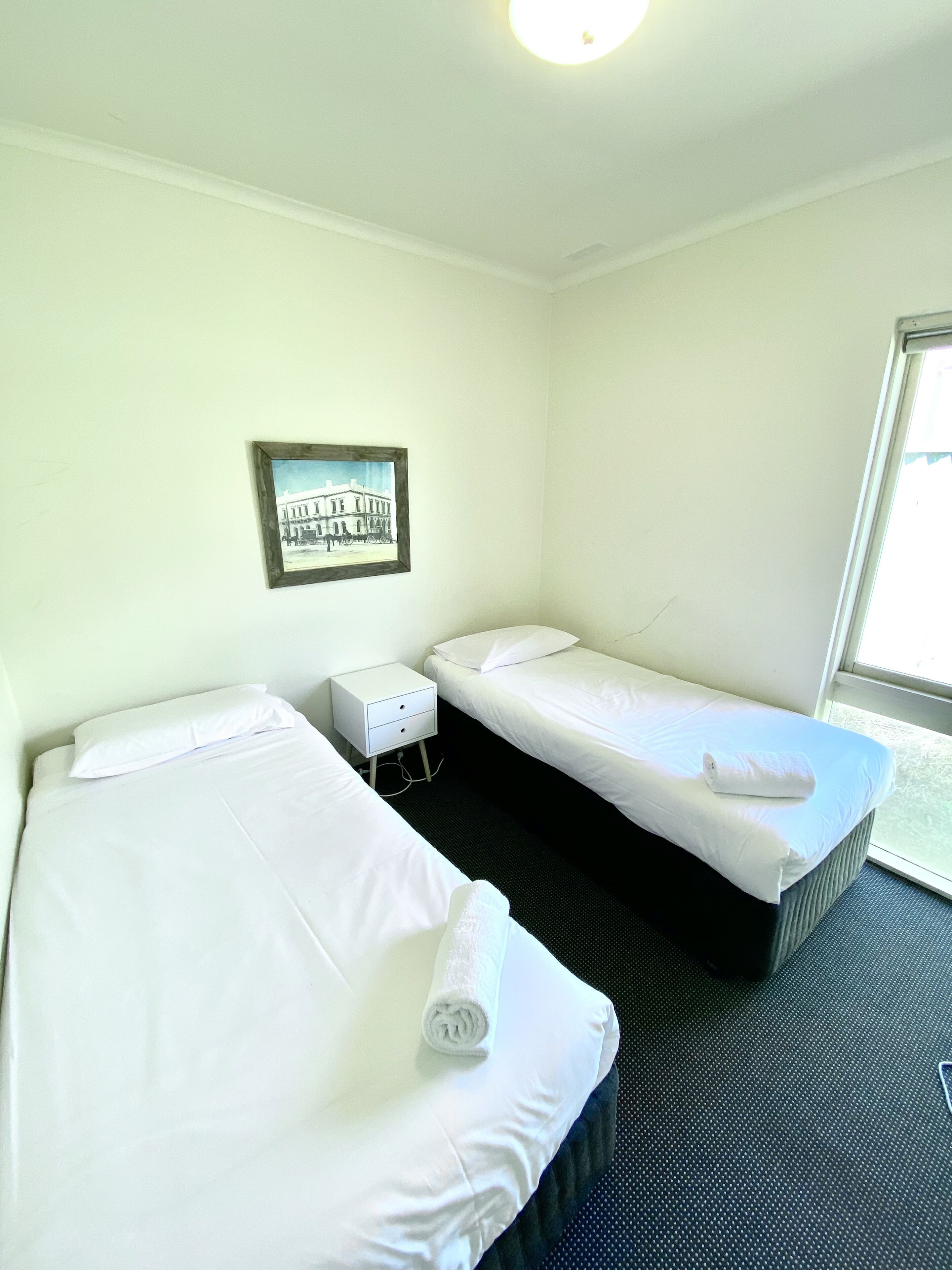 A hotel room with two single beds, a small bedside table with a lamp, a framed picture on the wall, and a window providing a view.