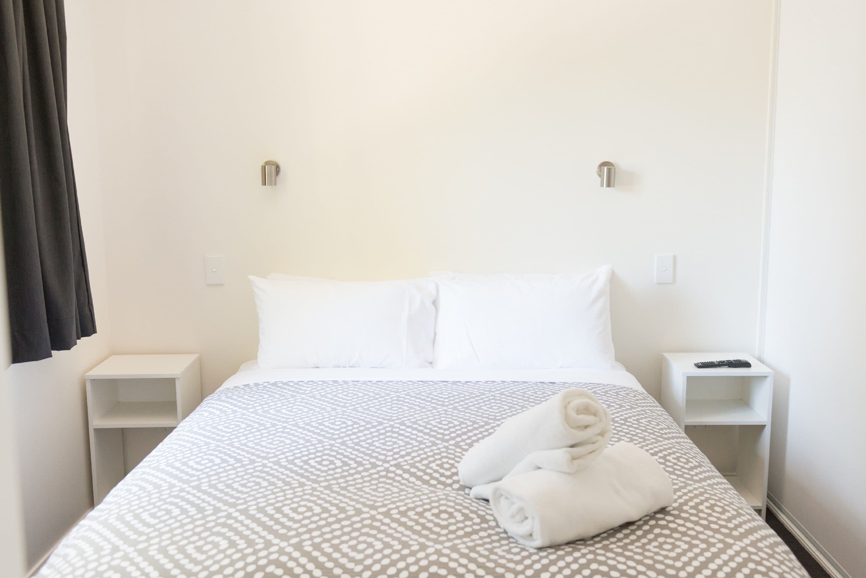 Quarto de hotel com uma cama, duas mesas de cabeceira e uma toalha dobrada sobre a cama.