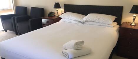 A neatly made bed with white linens, a rolled towel, and two bedside lamps. There are two chairs and a nightstand with a phone.