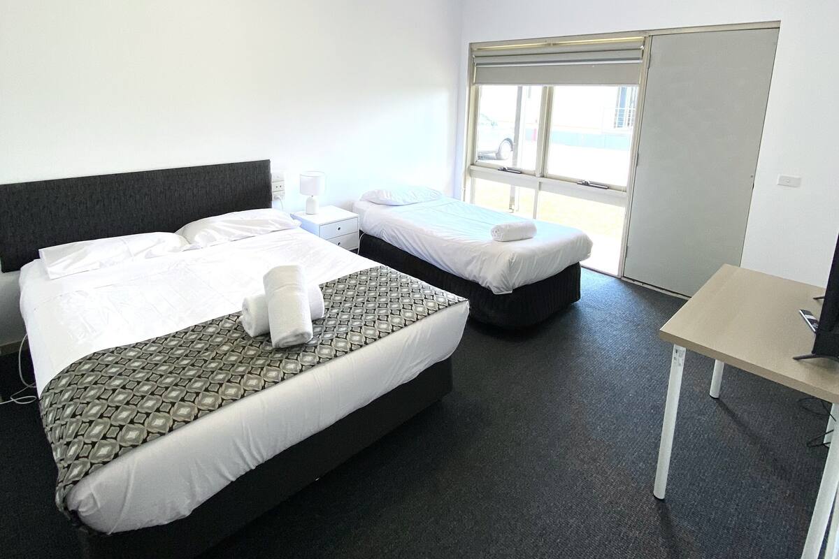 A hotel room with two beds, a desk, a window, and a door.