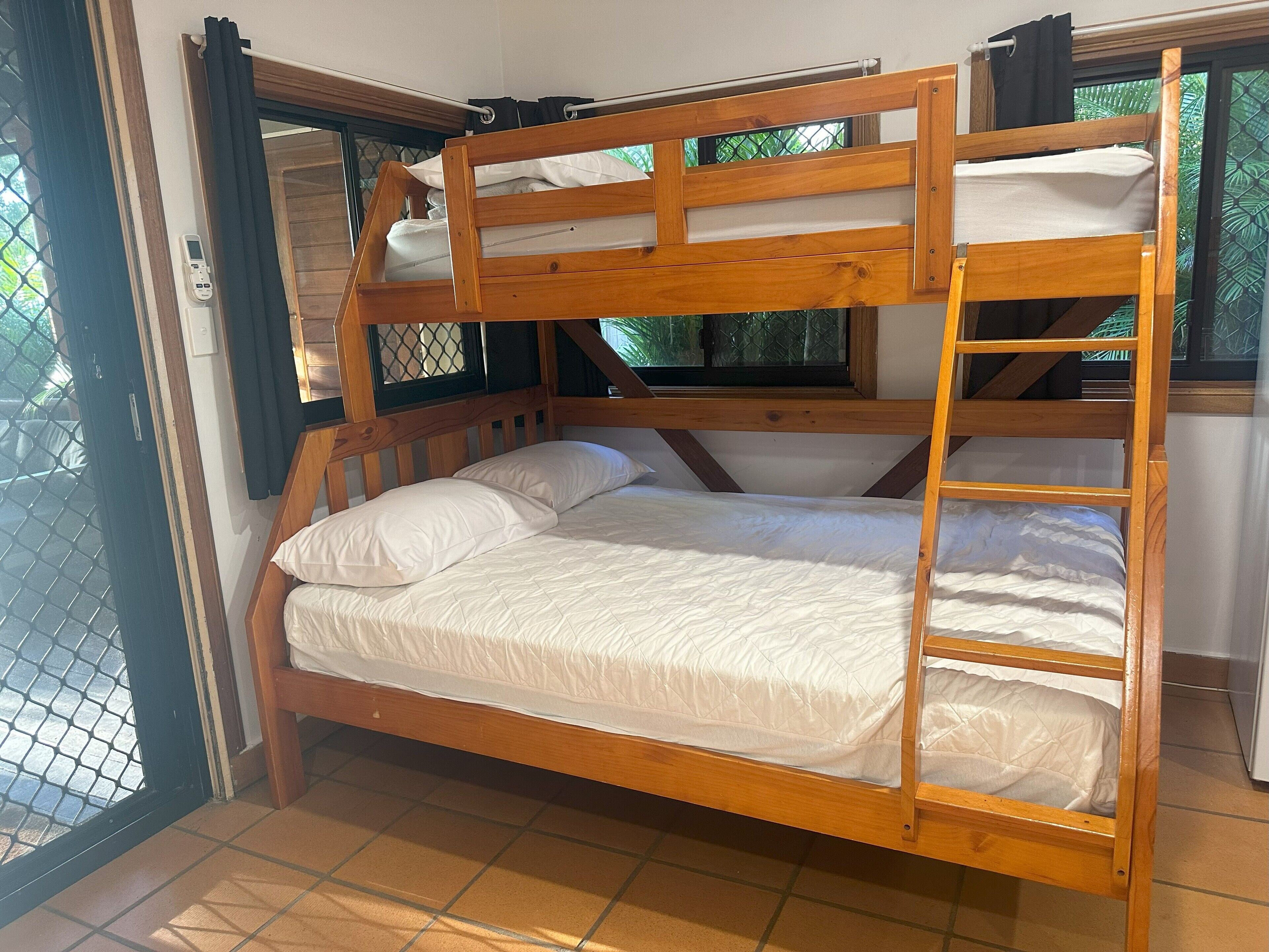 Una cama litera con estructura de madera, ropa de cama blanca y una ventana con vistas a follaje.