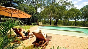 Zona de piscina amb gandules i una ombra, envoltada d'arbres i una tanca delimitant la piscina.