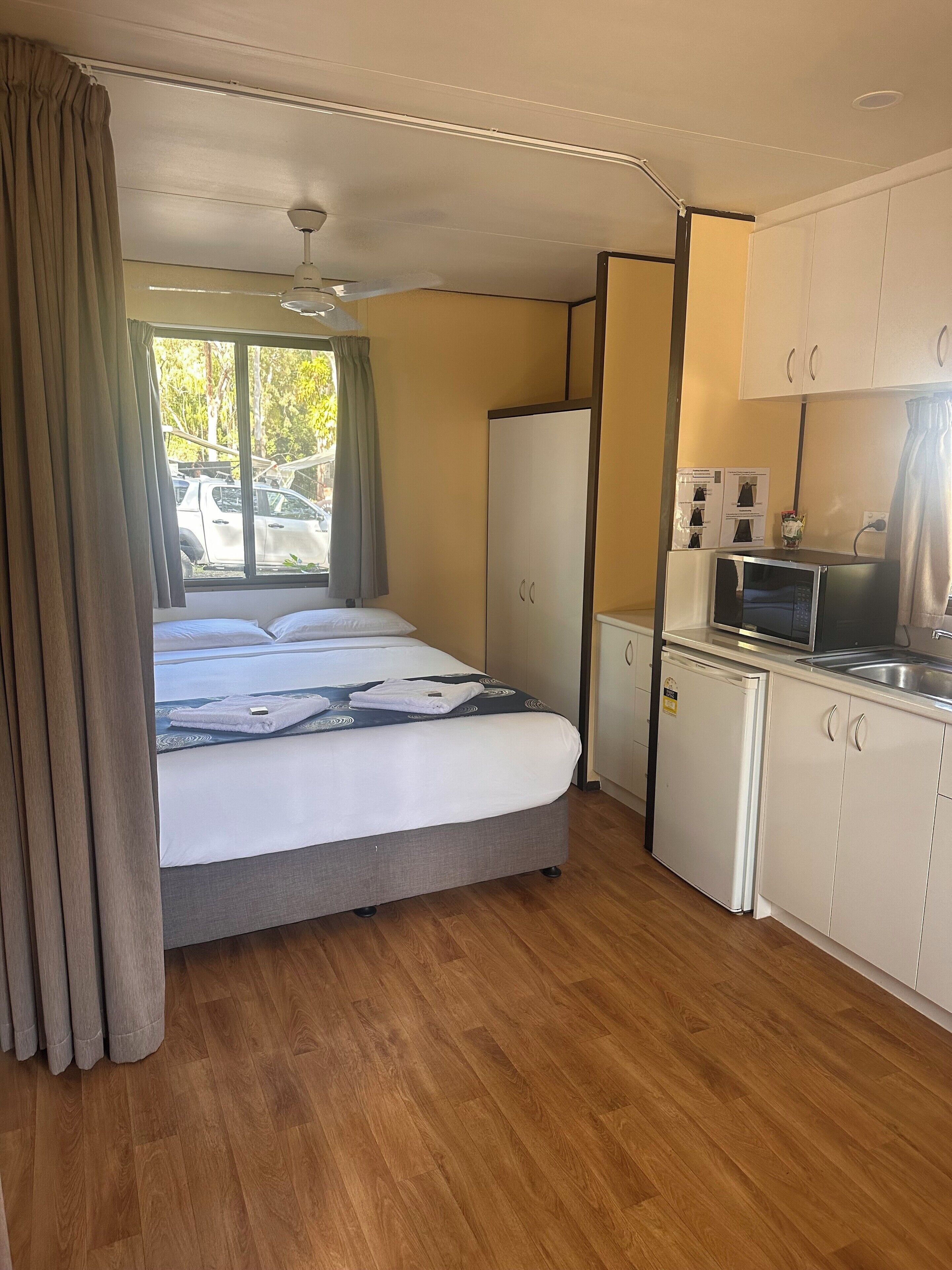 Une chambre avec un lit, un coin cuisine, un ventilateur de plafond et une fenêtre avec des rideaux.