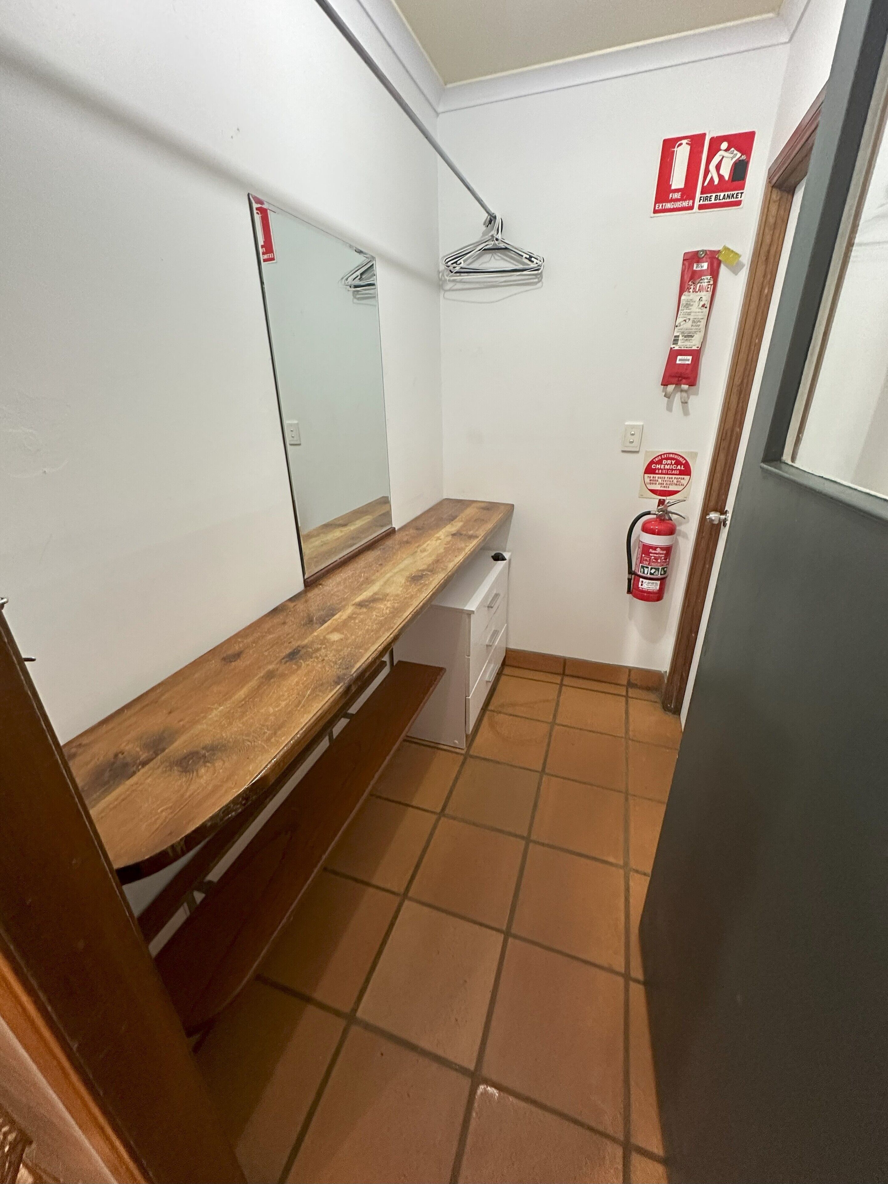 Una habitación pequeña y estrecha con un banco de madera, un armario blanco, un espejo y un extintor en la pared.