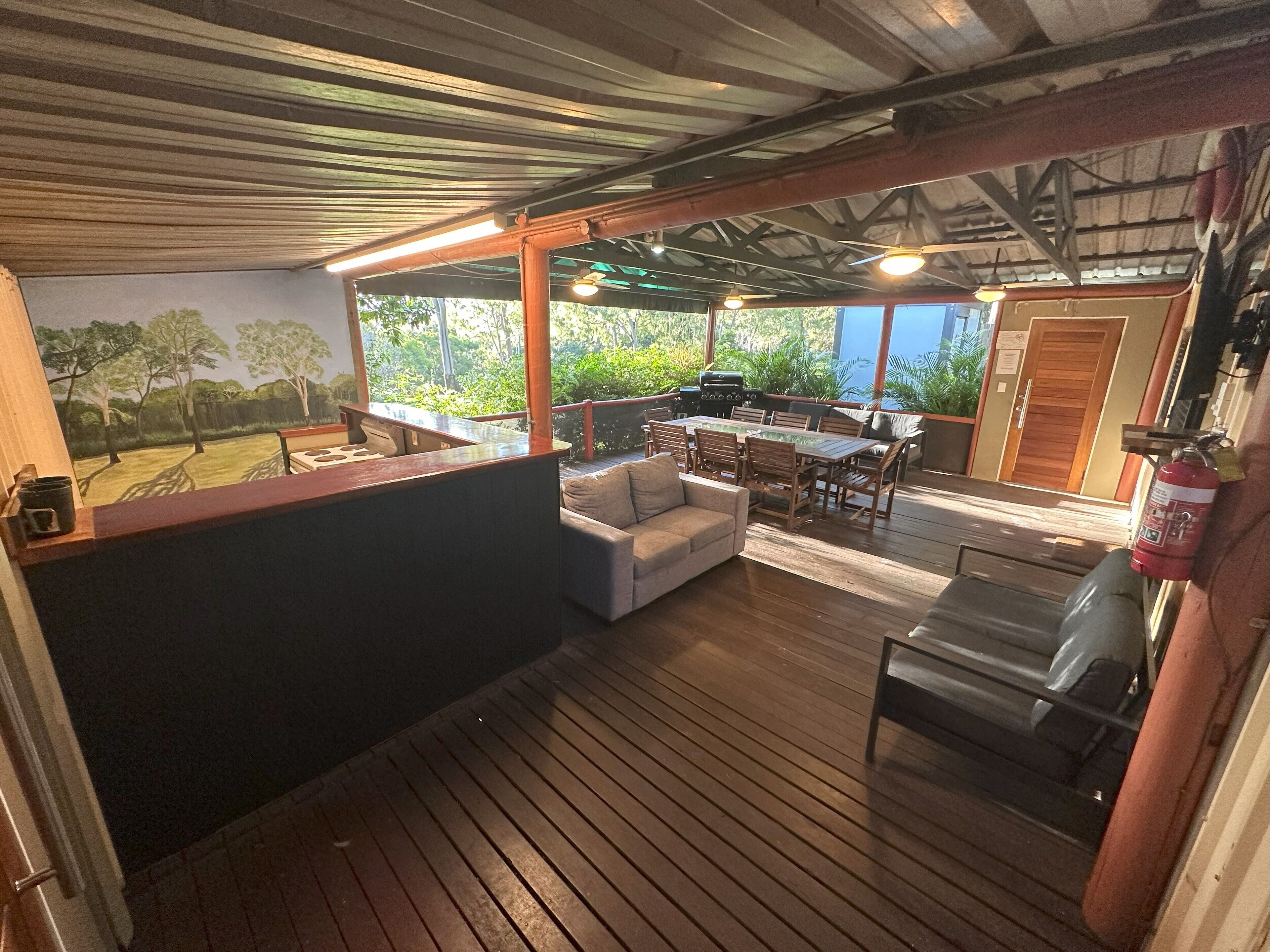 A covered outdoor area with a wooden deck, a barbecue grill, a dining table with chairs, and a sofa.