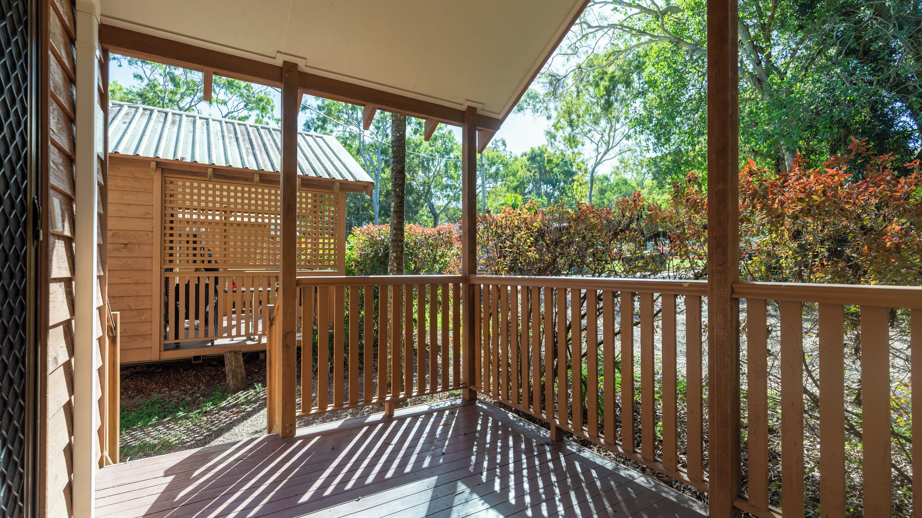 Una terraza de madera con un área cubierta, una pequeña construcción de madera, y un jardín con diversas plantas y árboles.