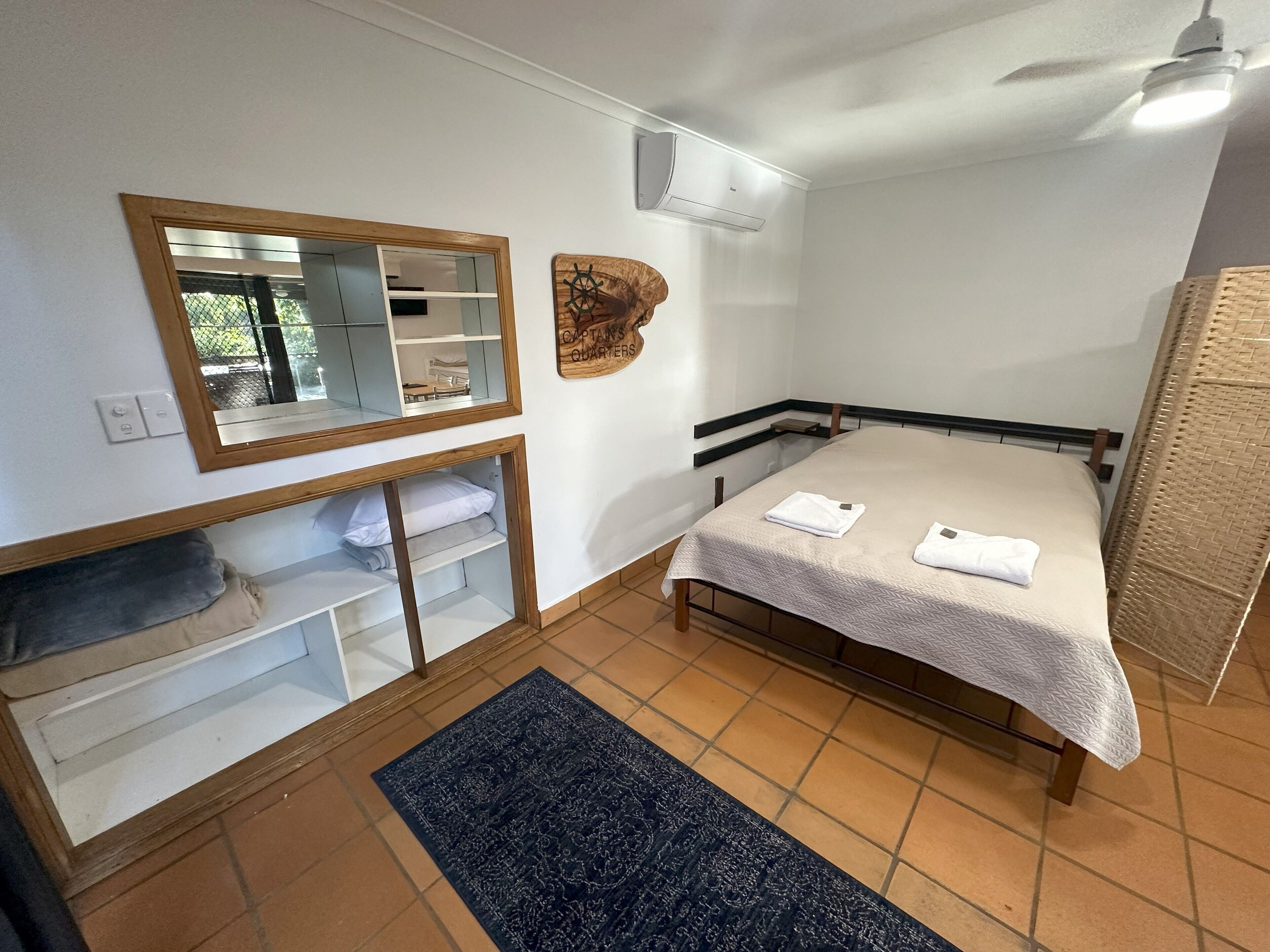A bedroom with a bed, a mirror, a wooden headboard, a small shelf, and a rug.
