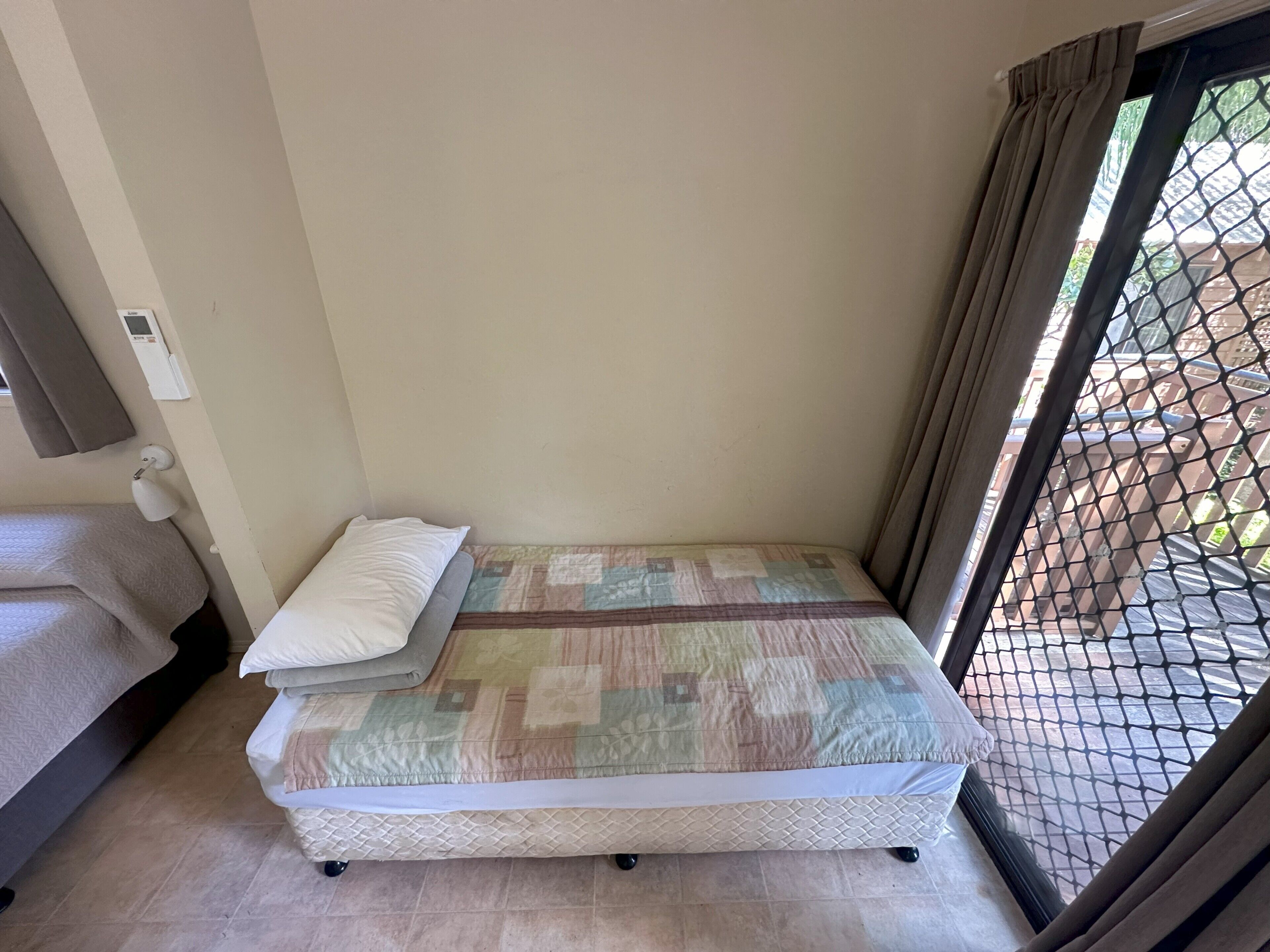 Une petite chambre avec un lit simple, un matelas matelassé, une lampe de chevet et une fenêtre donnant sur un bâtiment en briques.