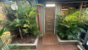 Una terrassa de fusta amb un camí central que condueix a una porta, envoltada de plantes verds exuberants i una paret de formigó.