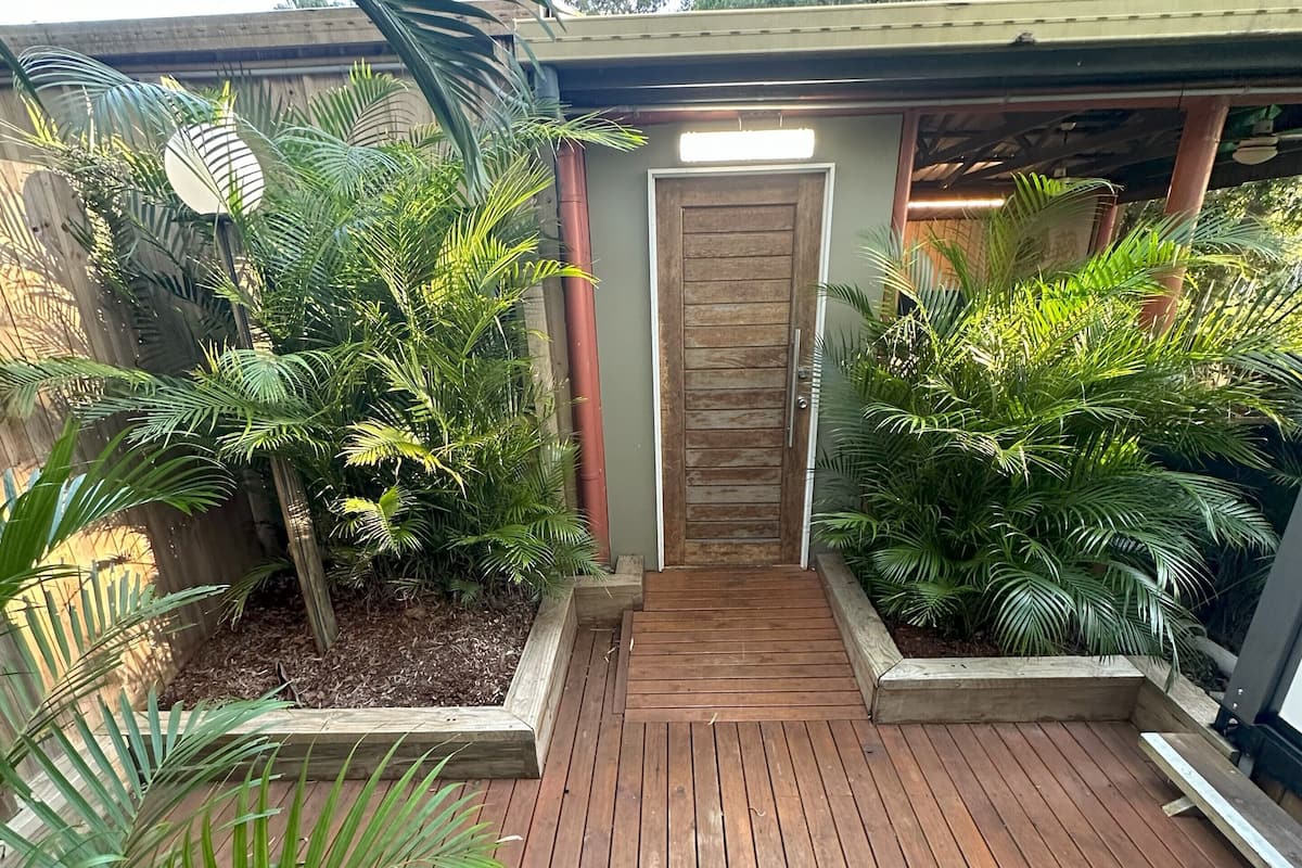 Une terrasse en bois avec un chemin central menant à une porte, entourée d’une végétation luxuriante et d’un mur en béton.