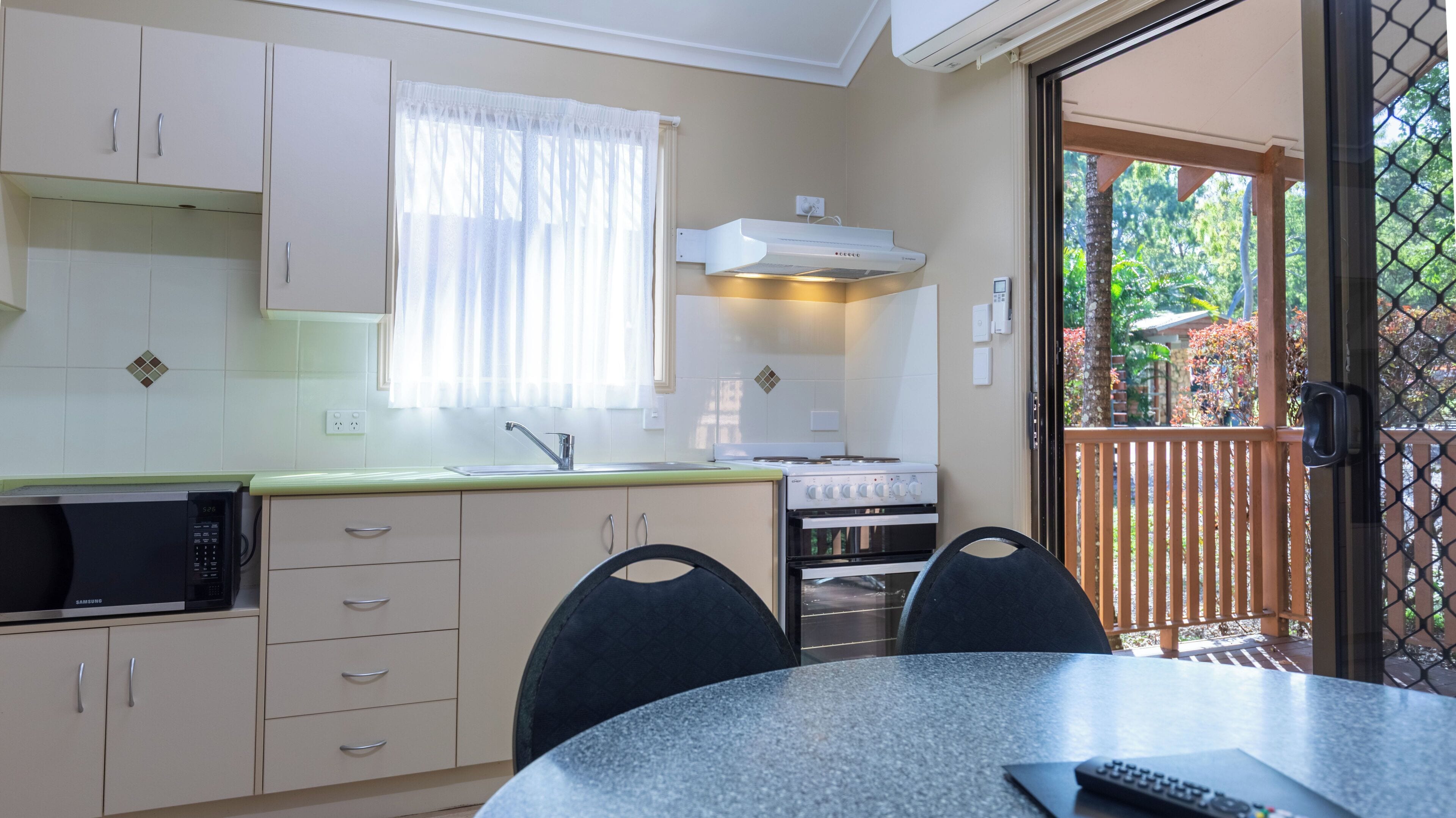 A compact kitchen with a microwave, stove, and sink, a dining table with chairs, and a view of the outdoors through a glass door.