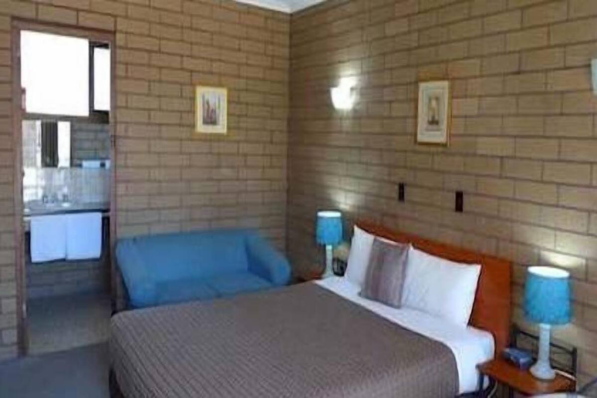 A hotel room with a brick wall, a bed with a brown headboard, two bedside lamps, and a view of a bathroom and kitchen area.