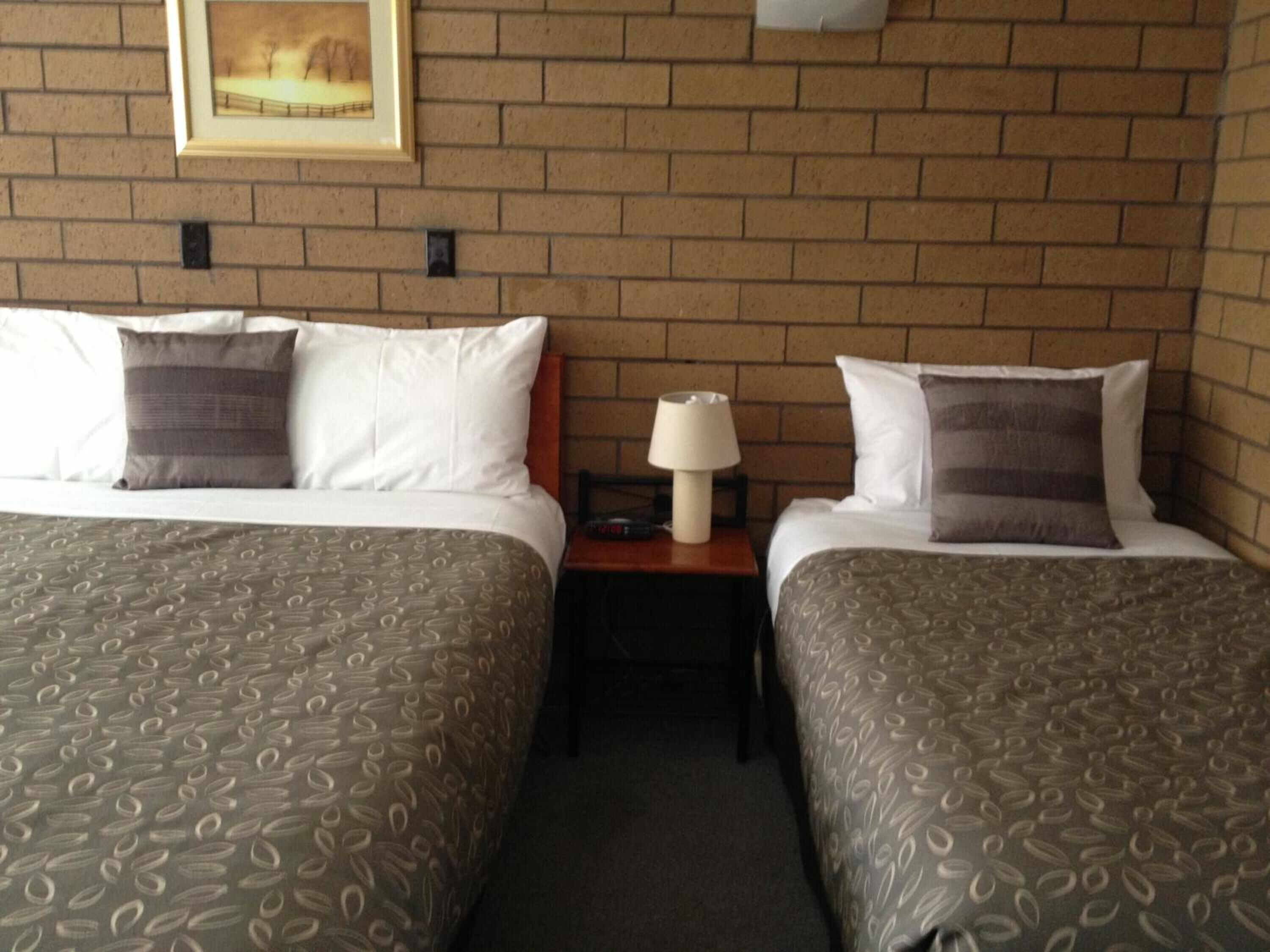 Habitación de hotel con dos camas, cada una con ropa de cama y almohadas estampadas, una mesita de noche con lámpara y un cuadro en la pared de ladrillo.