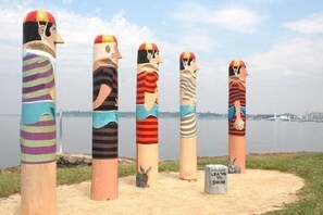 Une rangée de sculptures cylindriques hautes, ornées de motifs et de couleurs variés, est disposée sur une étendue de sable, avec un plan d’eau et le profil d’une ville en arrière-plan.