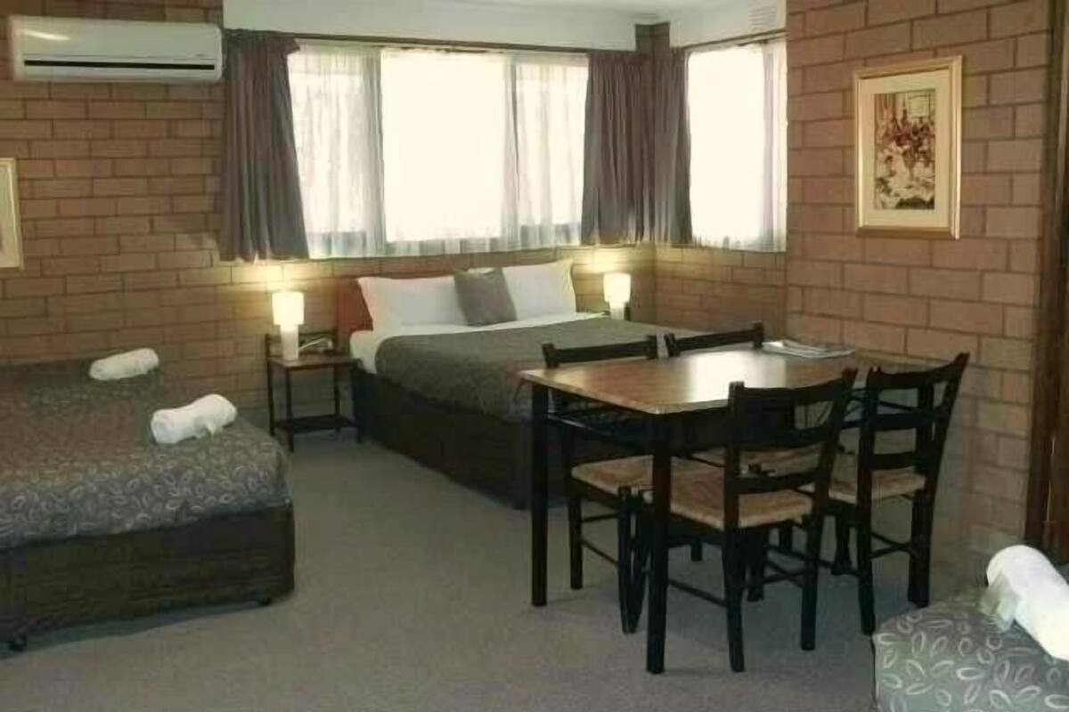 A hotel room with a bed, a small table, and two chairs. There is a window with curtains, an air conditioning unit, and a framed picture on the wall.