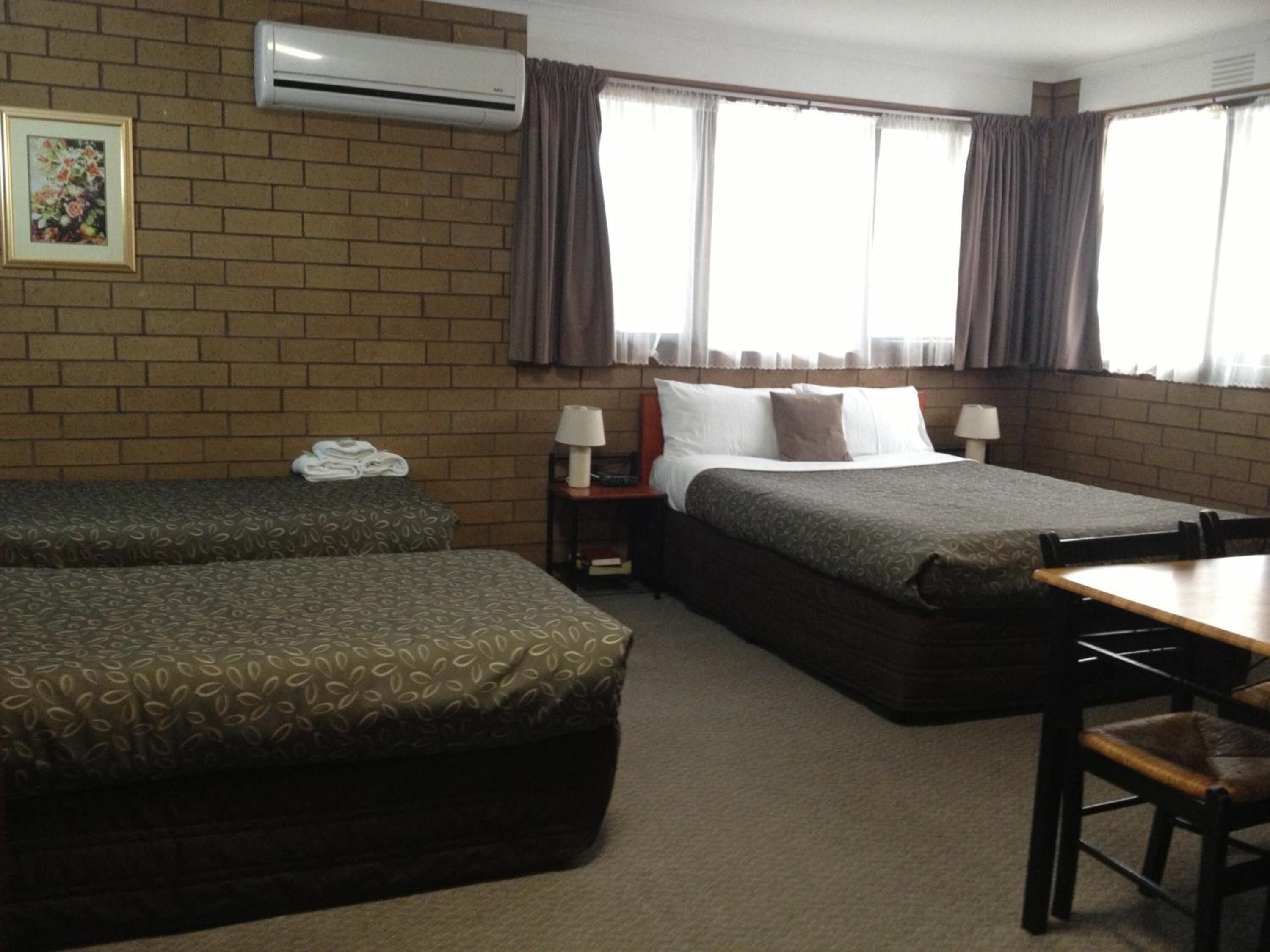 A hotel room with two beds, a desk, and a chair. There is a window with curtains, a wall-mounted air conditioner, and a framed picture on the wall.