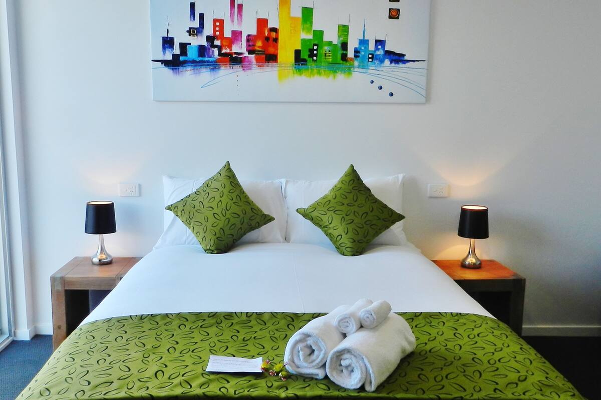 A hotel room with a neatly made bed, green patterned bedspread, two green pillows, two bedside lamps, and a colorful cityscape painting on the wall.