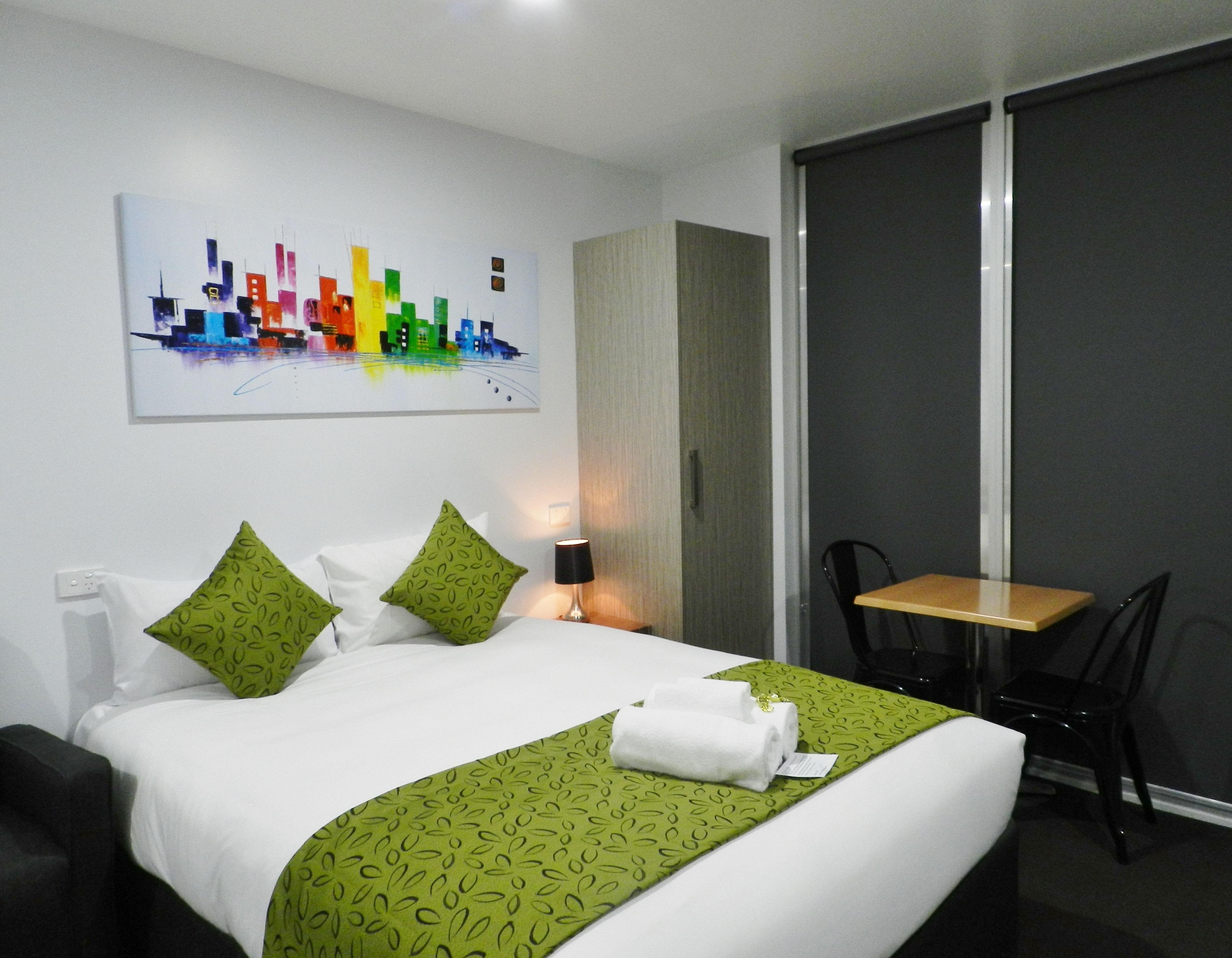 A hotel room with a bed, green patterned bedspread, white pillows, a cityscape painting on the wall, a desk with a chair, and a window with a view.