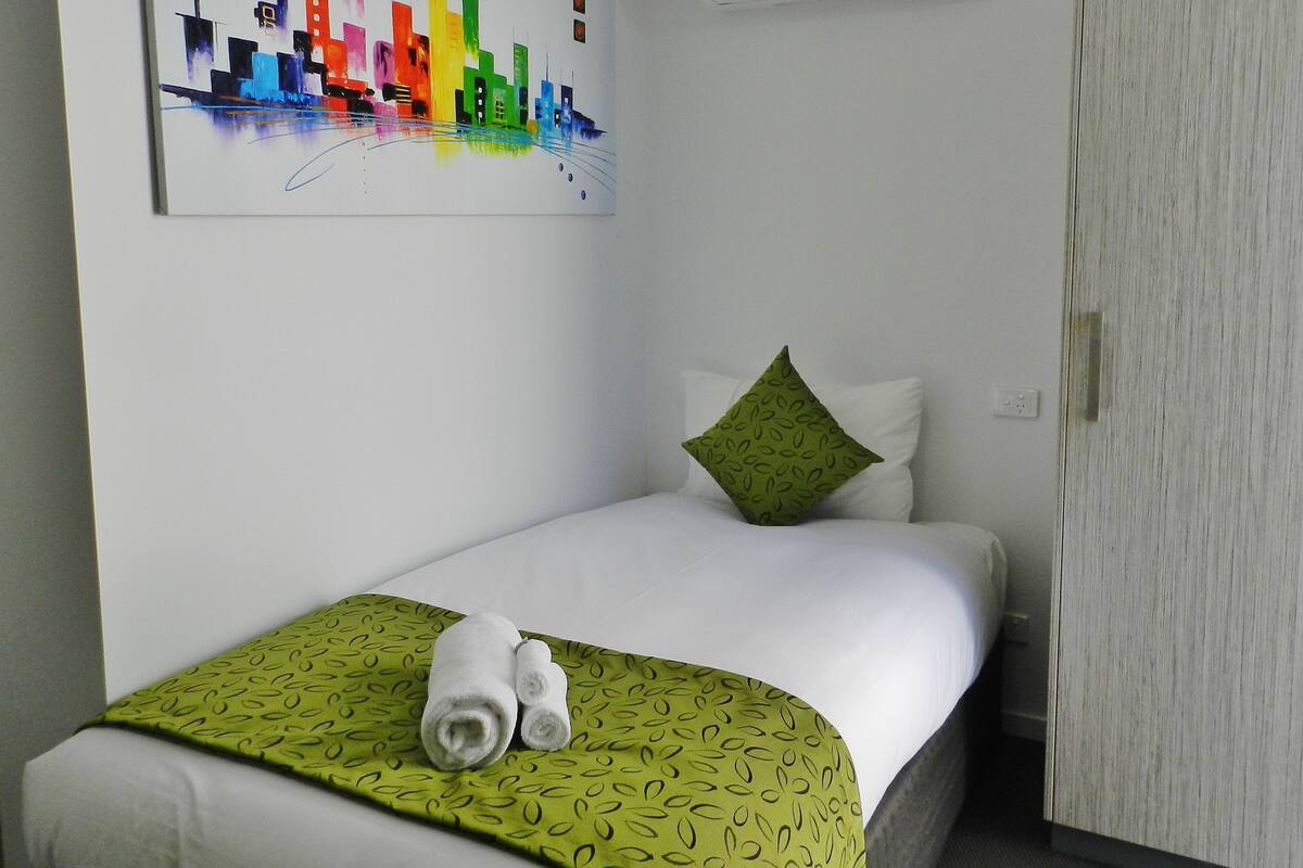 A hotel room with a bed, a green patterned throw, two rolled towels, a colorful abstract painting on the wall, and a white air conditioning unit.