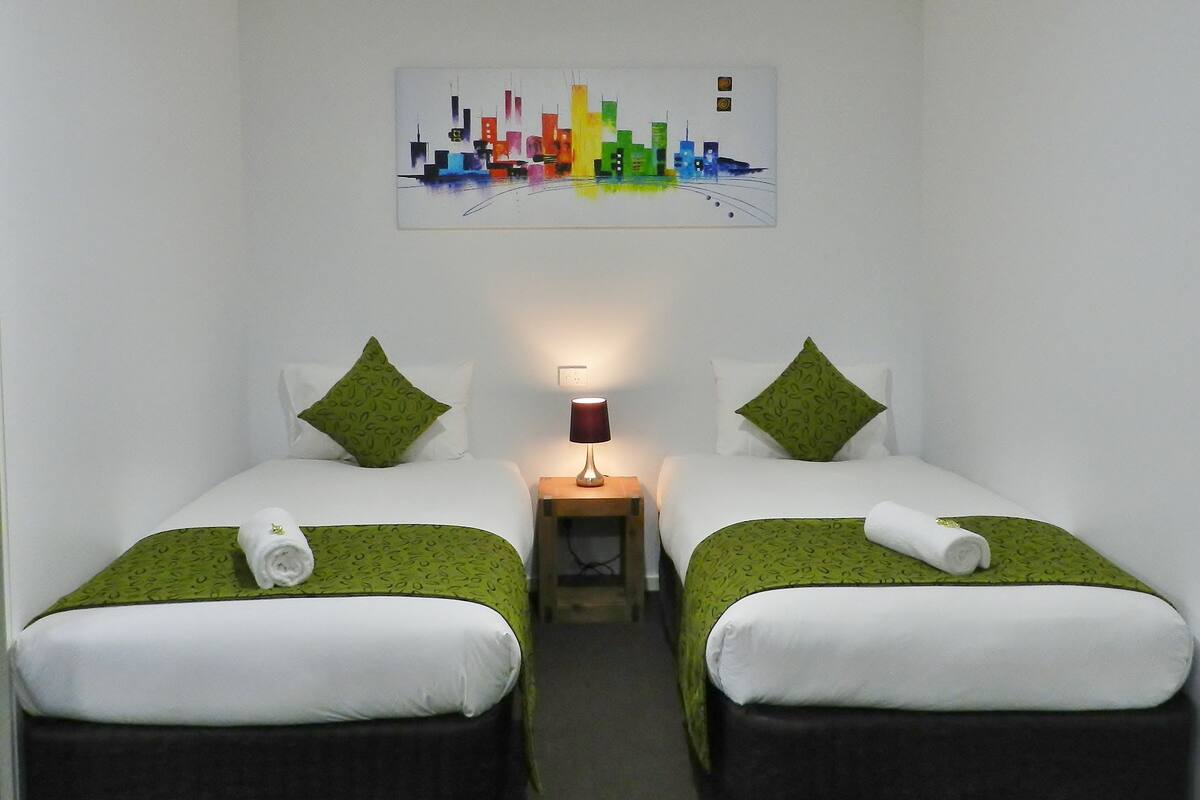 A hotel room with two beds, green patterned bedding, white pillows, a nightstand with a lamp, and a colorful cityscape painting on the wall.