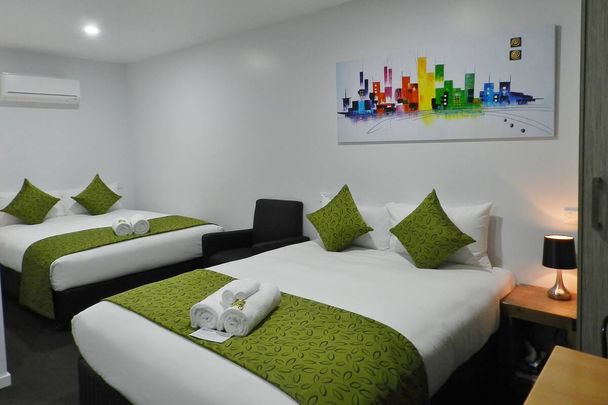 A hotel room with two beds, each with green patterned bedding and white pillows. There is a painting of a city skyline on the wall, a desk with a lamp, and an air conditioning unit on the wall.