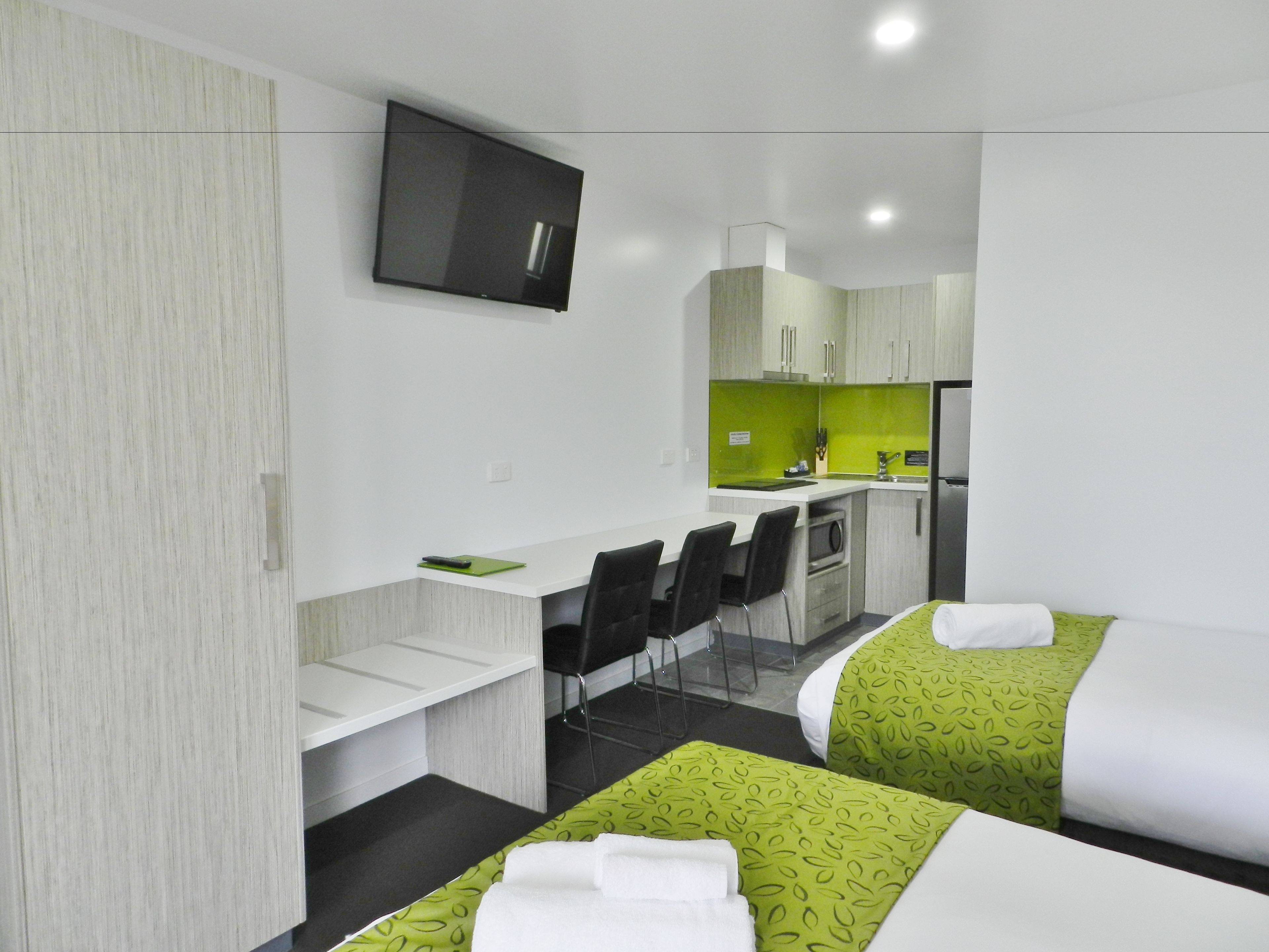 A modern hotel room with two single beds, a small desk, a chair, a TV, and a kitchenette.