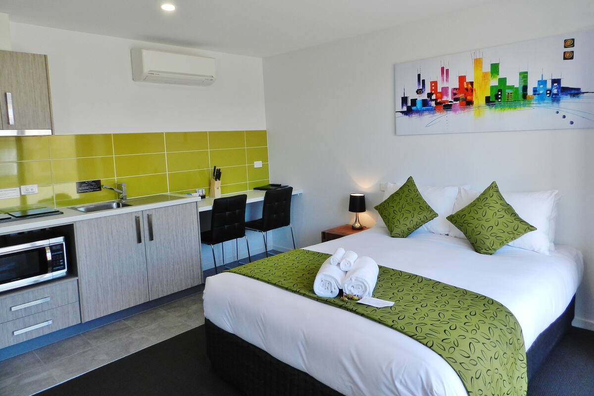 A hotel room with a bed, green patterned bedspread, two pillows, a nightstand with a lamp, a painting on the wall, a kitchenette with a microwave, and a green-tiled backsplash.
