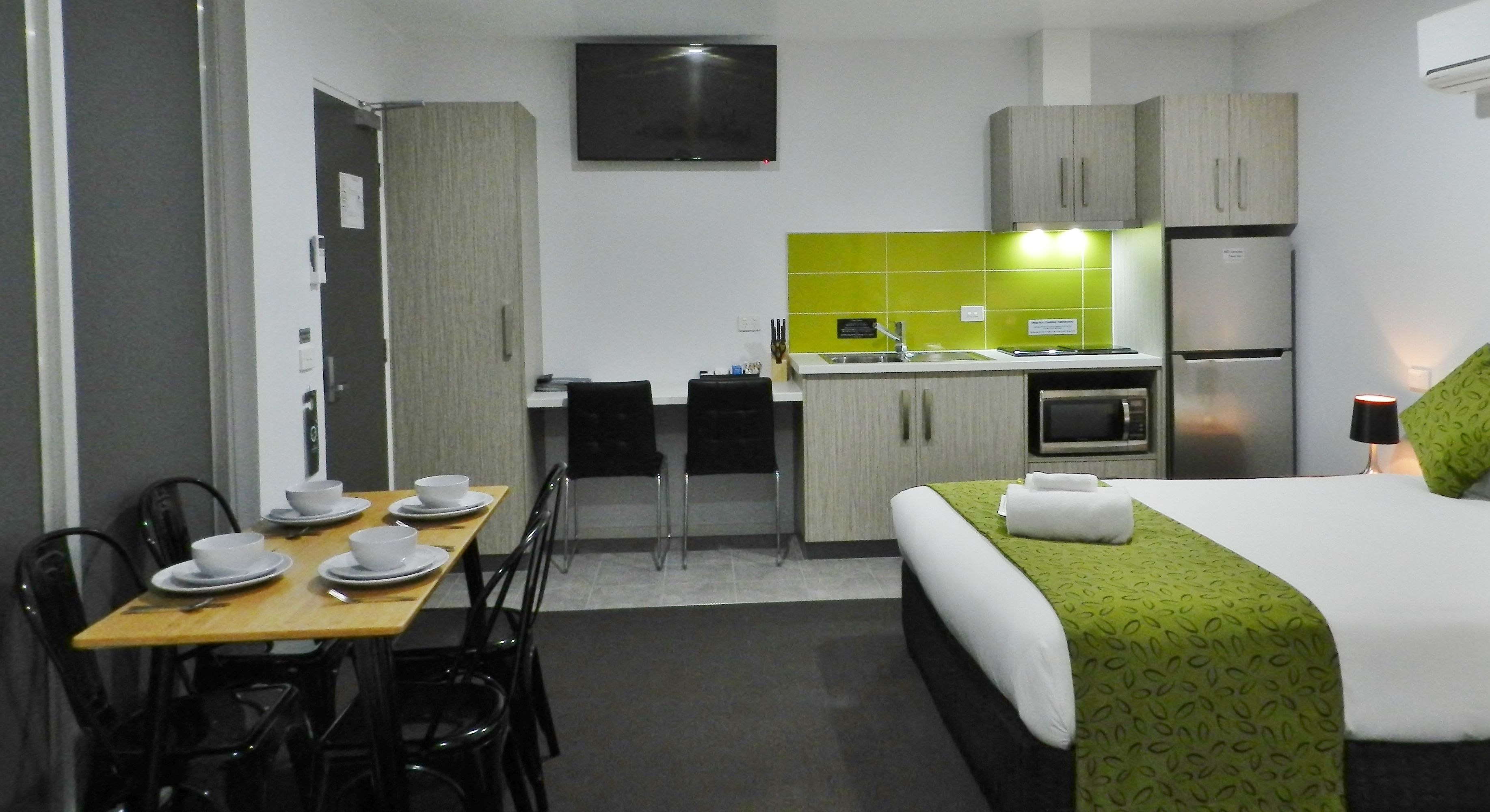 A hotel room with a bed, a dining table with chairs, a kitchen area with a sink and a refrigerator, and a television mounted on the wall.